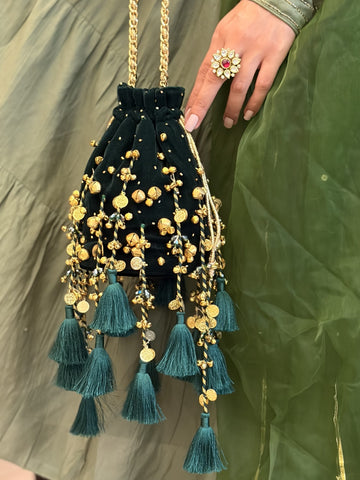 A green velvet drawstring potli bag decorated with gold coins, ghungroos, and tassels. Hand-embellished potli bag.